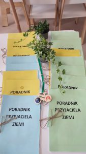 Na stole położone są obok siebie poradniki przyjaciela ziemi książeczka w kolorze niebieskim, żółtym, zielonym. Między nimi stoi roślinka w doniczce i medal w kształcie roweru.