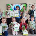 12 dzieci stoi na tle ekranu multimedialnego, na którym jest ziemia trzymana w rękach. Dzieci stoją obok siebie, w rękach trzymają doniczkę z roślinką, dyplom i książeczkę. Na szyi wisi medal w kształcie roweru.