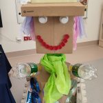 Na szarej podłodze stoi duży robot zrobiony z kartonów i odpadów plastikowych.