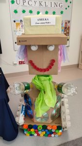Na szarej podłodze stoi duży robot zrobiony z kartonów i odpadów plastikowych.