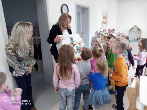Grupa dzieci patrzy na panią ubraną w czarną marynarkę, która trzyma duże pudło klocków.