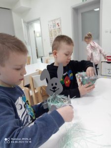 Dwóch chłopców w bluzie czarnej i niebieskiej wkładają sianko do koszyczka z zajączkiem.