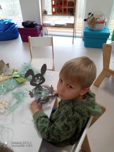 Chłopiec w bluzie zielonej wkłada sianko do koszyczka z zajączkiem.