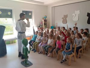 Dzieci siedzą na białych krzesłach i patrzą na pana Daniela obok którego stoi ufoludek. Pan Daniel ubrany jest w biały kombinezon kosmiczny.