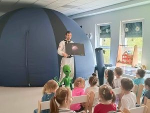 Dzieci siedzą na białych krzesłach i patrzą na pana Daniela obok którego stoi ufoludek. Pan Daniel ubrany jest w biały kombinezon kosmiczny i w ręce trzyma obraz planety.