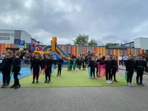 21 dzieci w czarnych strojach z kolorowymi chustami w pasie i na plecach tańczą na zielonym dywanie na tle tęczowych murów przedszkola.