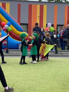 2 dziewczynki ubrane w strój żabki, tańczą na zielonym dywanie. Obok nich po bokach tańczą dzieci ubrane na czarno z kolorowymi chustami w pasie i na ramionach.