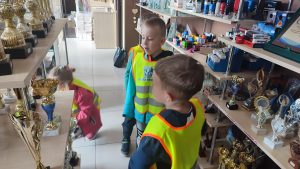 Dwóch chłopców ubranych w żółte kamizelki stoi obok regałów i patrzą na metalowe puchary i nagrody.