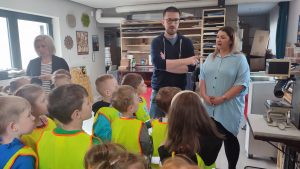 Duża grupa dzieci ubrana w żółte kamizelki stoją i słuchają panią i pana, którzy opowiadają o nakładaniu napisów na prezenty.