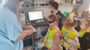 Dziewczynka w żółtej kamizelce i założonych czarnych okularach patrzy na laser, która graweruje napis na długopisie. Obok siedzi pan w okularach i granatowej bluzie.