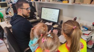 Kilka dziewczynek ubrane w żółte kamizelki bezpieczeństwa patrzą na ekran komputera, na którym pan w okularach robi wzory.