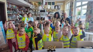 21 dzieci ubranych w kamizelki żółte bezpieczeństwa stoją na tle dużej liczby pucharów, nagród. Dzieci w rękach trzymają puchary, które podnoszą do góry. Za dziećmi stoją trzy kobiety.