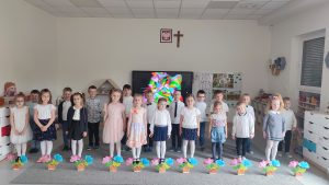 20 dzieci ubranych na galowo, stoją na szarym dywanie, przed nimi stoją kwiatki różowe i niebieskie w brązowych walizeczkach.