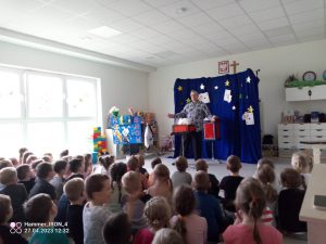 Grupa dzieci siedzi na szarym dywanie i patrzy na czarodzieja u branego w niebiesko białą bluzkę, który czaruje z czerwonych pudełek białe króliczki.