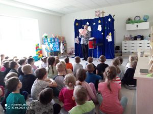 Grupa dzieci siedzi na szarym dywanie i patrzy na czarodzieja u branego w niebiesko białą bluzkę, który czaruje z dużych kart.