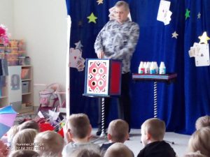 Grupa dzieci siedzi na szarym dywanie i patrzy na czarodzieja u branego w niebiesko białą bluzkę, który czaruje z czerwonego pudełka.