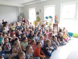 Duża grupa dzieci siedzi na szarym dywanie i ogląda pokaz magii.
