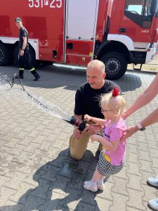Dziewczynka z pomocą strażaka trzyma wąż i próbuje odkręcać wodę jak prawdziwy strażak.