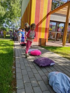 Dziewczynka w różowej piżamie biega slalomem na placu zabaw między poduszkami, w ręce niesie jajko.