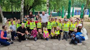 Duża grupa dzieci ubranych w kamizelki żółte - bezpieczeństwa stoją obok siebie i mają podniesione ręce do góry. Pośrodku dzieci stoi mężczyzna w niebieskiej bluzce.