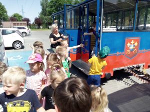 Grupa dzieci stoi przed kolejką, która jeździ po ulicach Ostrowa Wlkp. Kolejka jest w kolorach niebiesko-czerwonym.