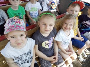 3 dziewczynki i 2 chłopców siedzi na drewnianej ławce i oglądają przez okno kolejki budynki Ostrowa Wlkp. Za nimi też siedzą dzieci.