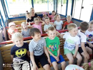 Na przodzie kolejki siedzi 5 chłopców ubrani w koszulki i spodenki na krótki rękaw. Za nimi siedzą pozostałe dzieci. Wszystkie dzieci są w środku kolejki.