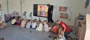 Dzieci siedzą na szarym dywanie i oglądają obrazy na tablicy interaktywnej.