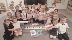 21 dzieci siedzi na szarym dywanie w strojach galowych. Przed nimi napis Dzień Starszaka.