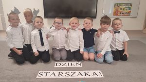 8 chłopców siedzi na szarym dywanie w strojach galowych. Przed nimi napis Dzień Starszaka.