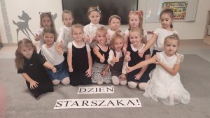 13 dziewczynek siedzi na szarym dywanie w strojach galowych. Przed nimi napis Dzień Starszaka.