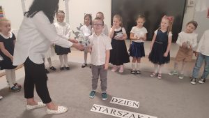 Chłopiec stoi na środku dywanu i otrzymuje prezent z rąk pani nauczycielki.