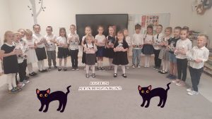 21 dzieci w strojach galowych stoją na szarym dywanie i trzymają w rękach prezenty. Przed nimi napis Dzień Starszaka.
