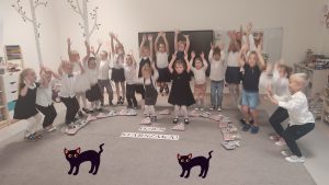 21 dzieci w strojach galowych stoją na szarym dywanie i podskakuje do góry. Przed nimi napis Dzień Starszaka.