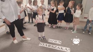 Dziewczynka stoi na środku dywanu i otrzymuje prezent z rąk pani nauczycielki.