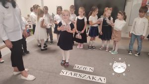Dziewczynka stoi na środku dywanu i otrzymuje prezent z rąk pani nauczycielki.