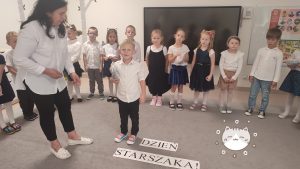 Chłopiec stoi na środku dywanu i otrzymuje prezent z rąk pani nauczycielki.