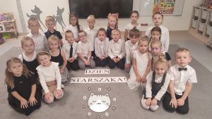 21 dzieci siedzi na szarym dywanie w strojach galowych. Przed nimi napis Dzień Starszaka.