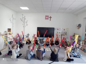 Dzieci siedzą na szarym dywanie i grają na kolorowych ,, bum bum rurkach".