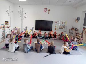 Dzieci siedzą na szarym dywanie i grają na kolorowych ,, bum bum rurkach".