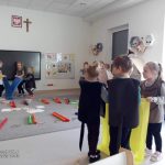 Grupki dzieci w szarej sali trzyma wstęgi materiału w różnych kolorach i rusza nimi do słyszanej muzyki.