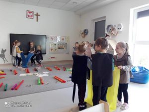 Grupki dzieci w szarej sali trzyma wstęgi materiału w różnych kolorach i rusza nimi do słyszanej muzyki.