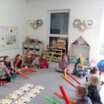 Dzieci siedzą na szarym dywanie i grają do muzyki na szarych ,,bum, bum rurkach".