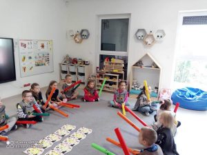 Dzieci siedzą na szarym dywanie i grają do muzyki na szarych ,,bum, bum rurkach".