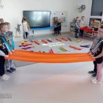 Grupki dzieci w szarej sali trzyma wstęgi materiału w różnych kolorach i rusza nimi do słyszanej muzyki.