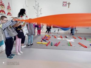 Grupki dzieci w szarej sali trzyma wstęgi materiału w różnych kolorach i rusza nimi do słyszanej muzyki.