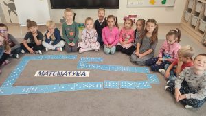 Dzieci siedzą w półkolu na szarym dywanie. Przed nim jest ułożony labirynt z klocków Domina z kropkami od 1 do 6.