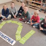 Chłopcy siedzą w półkolu na szarym dywanie. Przed nimi są ułożone cyfra: 2 i napis matematyka.