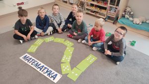 Chłopcy siedzą w półkolu na szarym dywanie. Przed nimi są ułożone cyfra: 2 i napis matematyka.