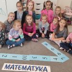 Dziewczynki siedzą w półkolu na szarym dywanie. Przed nimi są ułożone cyfry: 1 i napis matematyka.